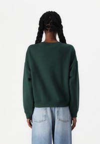 Tmavě zelená mikina s volným střihem, žebrovanými manžetami a kulatým výstřihem; v kombinaci se světle modrými širokými džíny.