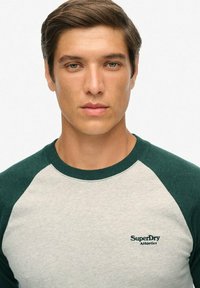 Ung mand med kort brunt hår iført en hvid og grøn raglan-t-shirt med "SuperDry Athletics" logo på brystet, der står med fronten vendt mod os.