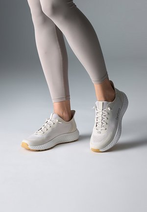 Personne portant des leggings beige et des baskets blanches à lacets avec semelles texturées, debout sur une surface grise lisse.
