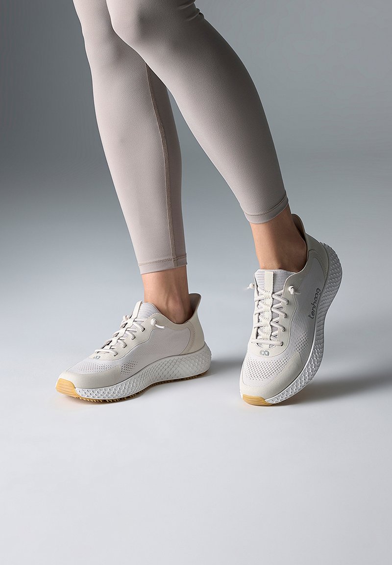 Person iført beige leggings og hvide snørestøvler med teksturerede såler, stående på en glat grå overflade.