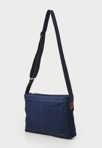 Tumši zila denim plecu soma ar kontrastējošām šuvēm. Iespējas: gara siksna, divas priekšējās kabatas un kvadrātveida forma.