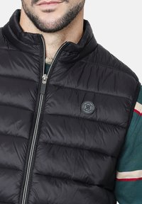 Chaleco acolchado negro con cuello alto, costuras horizontales y cierre con cremallera. Presenta un parche de logo redondo en el lado izquierdo del pecho.