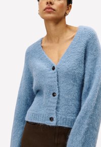 Frau, die eine hellblaue, fuzzy Strickjacke mit drei dunklen Knöpfen und braunen Hosen trägt, von Hals bis Hüfte gezeigt.