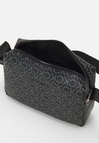 Czarna torba na ramię z materiału, z powtarzającym się wzorem logo CK, zamek błyskawiczny i odpinany pasek o strukturalnej fakturze. Wnętrze jest wyściełane.