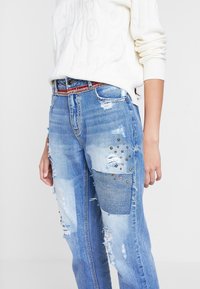 Jeans desgastados de color azul claro, con parches, tachuelas metálicas y una cinturilla texturizada de patrón rojo y blanco. Corte relajado en general.