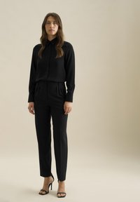 Zwarte button-up shirt en op maat gemaakte broek, beide gemaakt van een gladde stof. Slim fit met een subtiele glans, gecombineerd met zwarte hoge hakken sandalen.