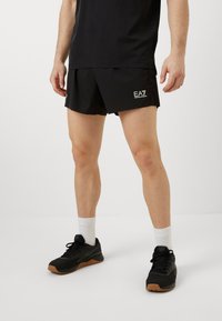 Μαύρες αθλητικές βερμούδες με λογότυπο στο στρίφωμα, συνδυασμένες με μαύρο μπλουζάκι και μαύρα αθλητικά παπούτσια με καουτσούκ �σόλα και λευκές κάλτσες.