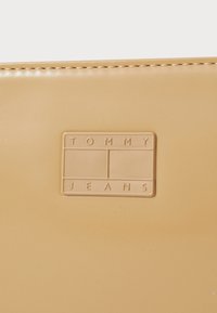 Beige syntetisk materialekortholder med preget "TOMMY JEANS"-logo og rektangulært design med jevn tekstur og sømmet kanter.