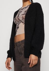 Žena v černém huňatém kardiganu přes květinový crop top s vysokým pasem a tmavě hnědými džínsy, stojící na jednolitém pozadí.