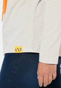 Beige långärmad skjorta med en orange grafisk design, försedd med en nederkantsetikett i orange med en svart oändlighetssymbol. Bärs tillsammans med mörkblå jeans.