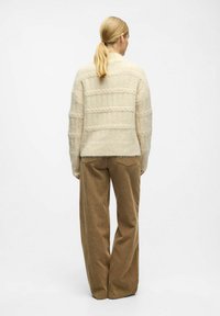 Kobieta z blond włosami w kucyku, ubrana w sweter w kremowym kolorze z dzianiny oraz brązowe szerokie spodnie z korduroy, stojąca na tle białej ściany.