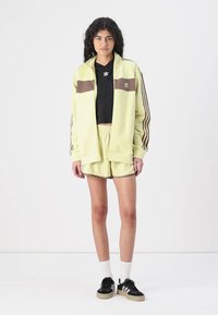 Light yellow tracksuit jacket with brown accents, paired with a black top and matching yellow shorts. Black sneakers with white socks.