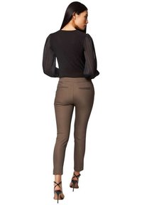 Bodysuit noir avec des manches longues plissées et transparentes, associé à un pantalon marron fuselé. Des chaussures à talons mettent en valeur la silhouette élégante de la tenue.