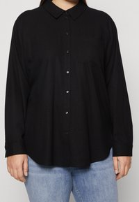 Schwarzes Hemd mit Knopfleiste, Kragen, langen Ärmeln und einer einzelnen Brusttasche. Aus weichem Stoff gefertigt, mit glänzenden schwarzen Knöpfen.
