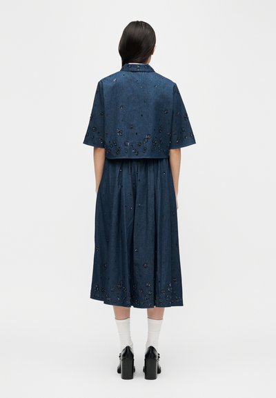 Femme aux cheveux foncés vue de dos, portant une tenue deux pièces bleu foncé ornée de motifs floraux, des chaussettes blanches et des chaussures à talons noires.