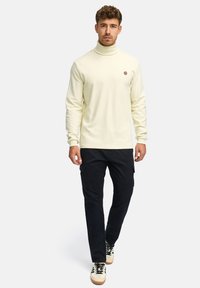 Pull col roulé crème avec manches longues, en tissu texturé, et petit patch logo, associé à un pantalon cargo foncé et des baskets.
