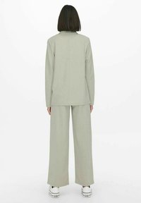 Lichtgrijs, op maat gemaakt pak bestaande uit een lange mouwen jasje en wijde broekspijpen, gemaakt van soepele stof, gedragen met witte sneakers.