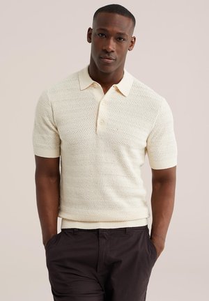 Homme portant un polo en maille crème à manches courtes avec un motif en zigzag texturé et un pantalon foncé, debout les mains dans les poches.