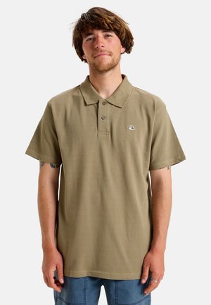 Jonge man met bruin haar, gekleed in een olijfgroen poloshirt met korte mouwen en een blauwe broek, staand tegen een effen achtergrond.