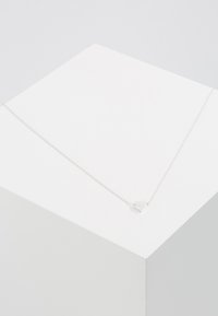 Collier en chaîne argentée avec un pendentif en forme de cœur au fini poli, exposé sur un piédestal blanc. Design simple, épaisseur moyenne.