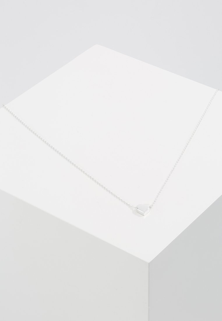 Collier en chaîne argentée avec un pendentif en forme de cœur au fini poli, exposé sur un piédestal blanc. Design simple, épaisseur moyenne.