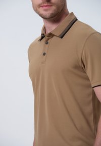 Polo color caqui con un cuello con acentos negros, tapeta de tres botones y una tela suave y texturizada.