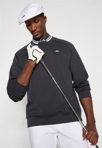 Sötétszürke pulóver kerek nyakkivágással és fehér logóval, fehér sapkával, fekete logóval, golfütőt tartva, fehér kesztyűben.