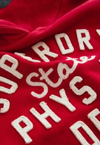 Röd bomullshoodie med texturerade krämfärgade broderade bokstäver som stavar "Superdry" och "Physical Education." Uppmärksammade stygn detaljer finns.