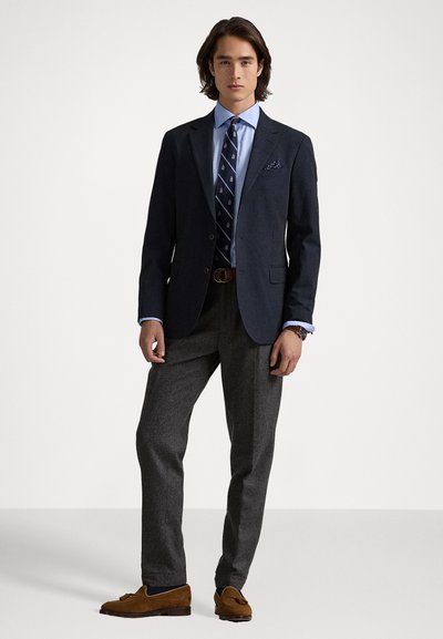 Blazer marine sur chemise bleu clair, cravate à motif, pantalon gris, chaussures en daim marron ; comporte un pochette et une ceinture en cuir.