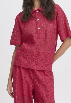 Kvinde iført en rød blomsterblonde kortærmet skjorte med perleknapper og matchende blondebukser mod en ensfarvet baggrund.