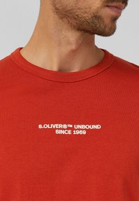 Rode katoenen T-shirt met ronde hals, korte mouwen en witte opdruk met de tekst "s.OLIVER® UNBOUND SINCE 1969" over de borst.