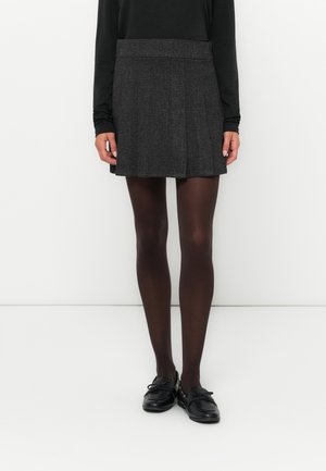 Kvinde iført en mørkegrå plisseret mininederdel, sorte gennemsigtige strømpebukser, sort langærmet top og sorte loafers, stående mod en hvid baggrund.