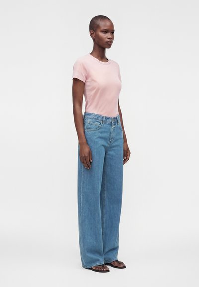 Personne de profil portant un t-shirt rose pâle à manches courtes, un jean bleu à jambes larges et des sandales noires sur fond blanc uni.