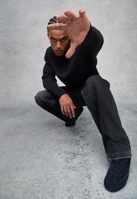 Schwarzer gerippter Strickpullover, dunkelgraue Jeans und schwarze Wildledersneaker. Hände nach vorne ausgestreckt in einer hockenden Position vor einem neutralen Hintergrund.
