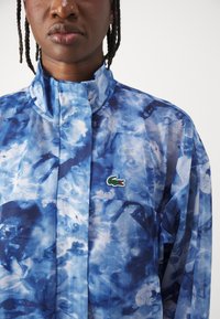 Modrá bunda s abstraktním vzorem vyrobená z lehkého, průhledného materiálu. Má vysoký límec a malé zelené logo Lacoste na hrudi.