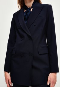 Navy double-breasted blazer in textured fabric, featuring a notched collar, two front pockets, and button accents. Scarf tied at the neck.