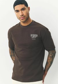 Brun kortærmet t-shirt med rund hals, der har hvid tekst "San Francisco California" og et minimalistisk grafisk motiv på venstre bryst.