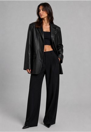 Woman wearing a black leather blazer, black crop top, wide-leg black trousers, and black pointed shoes standing against a gray background.
