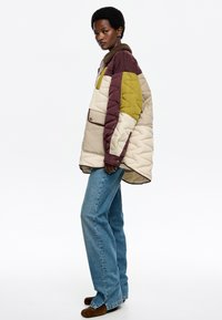 Chaqueta acolchada en beige, marrón y verde oliva. Presenta un cierre de botones, un cuello y un corte relajado. Combinada con vaqueros azules y zapatos marrones.