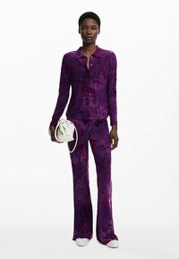 Lilla sametist komplekt, mis koosneb pikkade varrukatega nööbitavast särkist ja lehvikpükstest, millel on lillemustri ning mida kannab valge käekott.