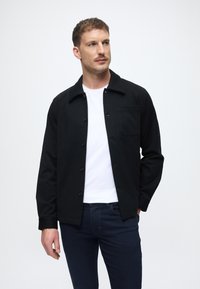 Čierna blúzka na gombíky s golierom, s vreckom na hrudi, nosená na bielom tričku a tmavých džínsoch. Hladký povrch látky.