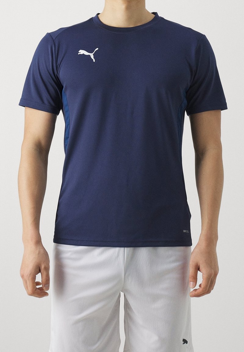 Mand iført navyblå Puma kortærmet sportstrøje og hvide shorts, stående med armene afslappet ned langs siden.