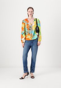 Blouse colorée à manches longues avec des motifs floraux et un décolleté en V, associée à un jean bleu avec des accents floraux brodés à l'ourlet.