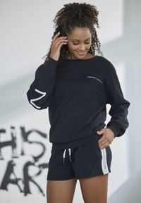 Black cotton sweatshirt with long sleeves and navy shorts, featuring a white logo on the front and accented drawstrings in light blue.