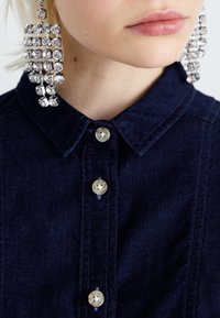 Camicia di jeans blu scuro con colletto a punta e tre bottoni color crema. Accompagnata da grandi orecchini a forma rettangolare in cristallo.