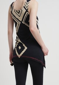 Musta hihaton paita, jossa on vaaleanbeige geometrinen kuvio, korkea-alhainen helma ja punaiset ompelutiedot. Valmistettu kevyestä kankaasta.