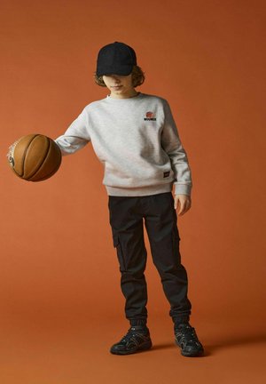 Sudadera gris con el logo "BOUNCE", pantalones cargo negros y zapatillas negras. Sosteniendo un balón de baloncesto, con una gorra negra. Fondo naranja.