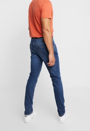 Hombre con jeans ajustados azules, camisa naranja de manga corta y zapatillas blancas Asics, de pie de lado contra un fondo liso.