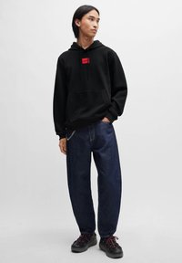 Svart hettegenser med en rød logo-lapp, kombinert med løse mørkeblå jeans og klumpete svarte sko med røde snørelisser.