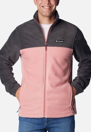 Mann in einer zweifarbigen Fleecejacke mit dunkelgrauem Oberteil und Ärmeln sowie pinkem Unterkörper, der mit den Händen in den Taschen dasteht.
